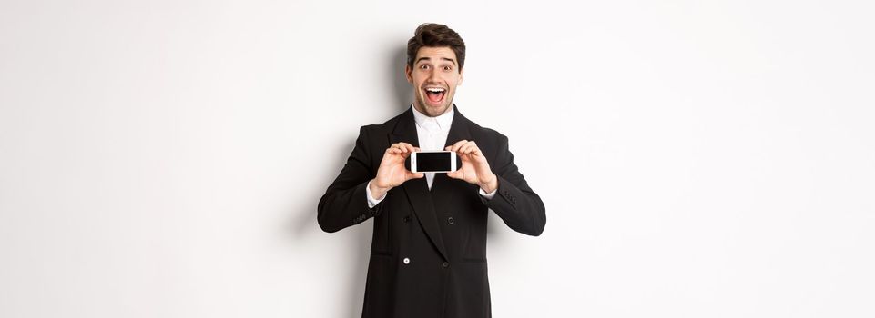 Image of cheerful, handsome man in black suit, showing smarthone screen and looking amazed, standing against white background.