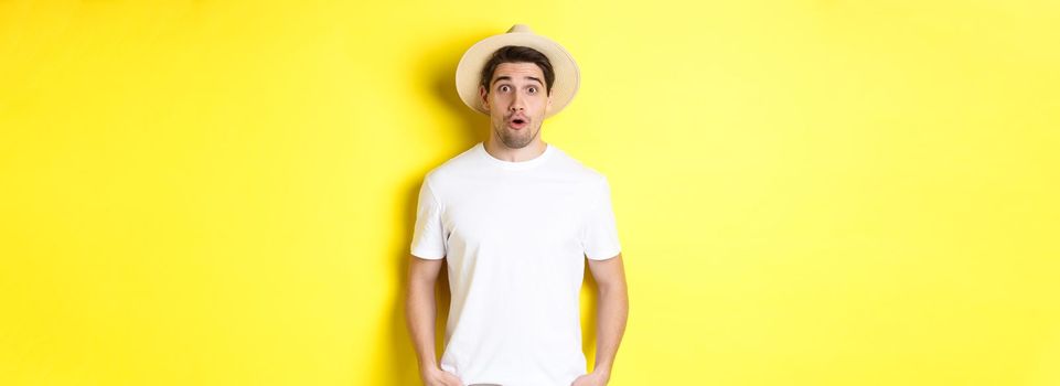 Concept of travelling and tourism. Surprised caucasian guy in straw hat, say wow, standing against yellow background.