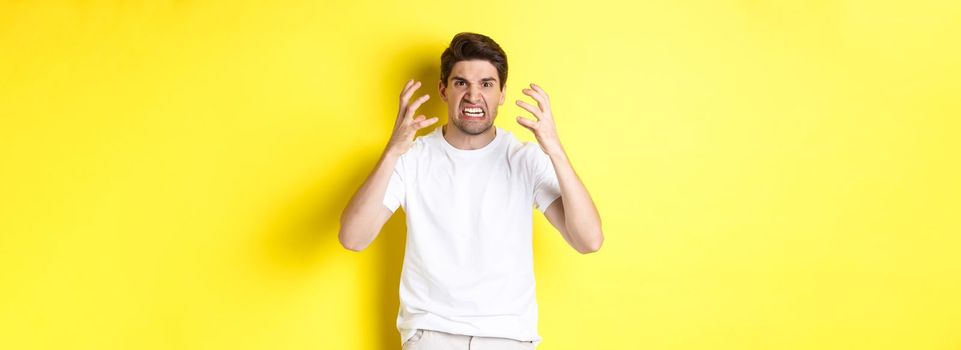 Angry man looking mad, grimacing and shaking hands furious, standing outraged against yellow background.
