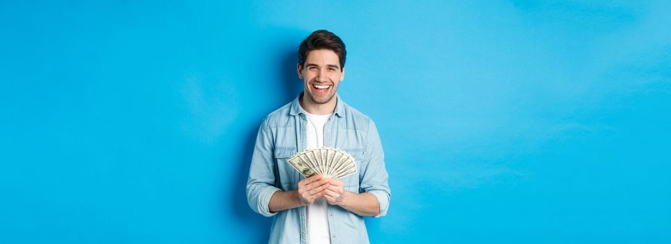 Happy successful man smiling pleased, holding money, standing over blue background.