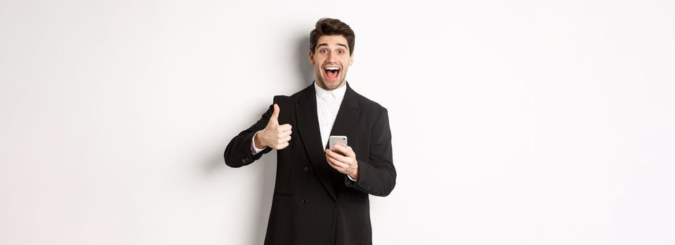 Portrait of handsome man in trendy suit showing thumbs-up in approval, using mobile phone app, smiling pleased, standing over white background.