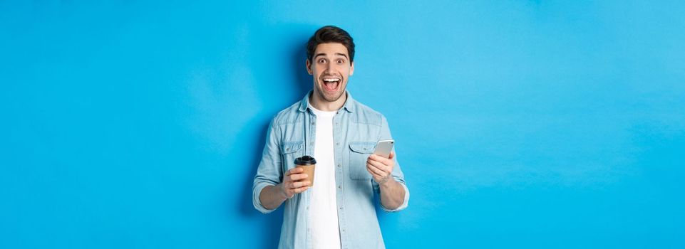 Happy young man drinking coffee and using mobile phone, looking excited, standing against blue background.