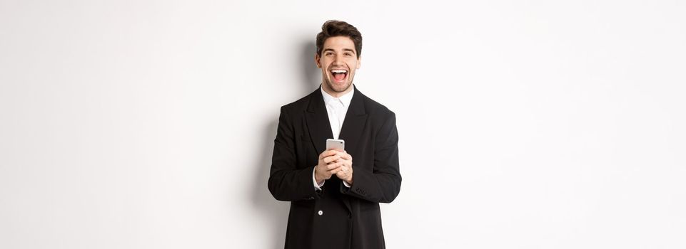 Portrait of happy good-looking man, wearing black suit, laughing from happiness and using mobile phone, standing over white background.