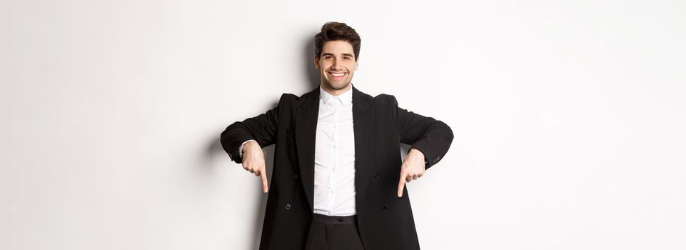 Portrait of good-looking stylish man in black suit, pointing fingers down and smiling, showing winter holidays promo, standing over white background.