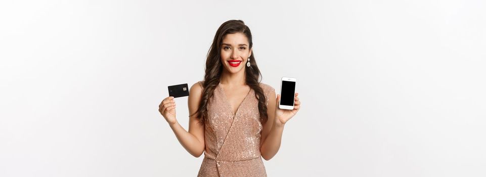 Online shopping and holidays concept. Attractive woman in party dress showing credit card and mobile screen, smiling happy, white background.