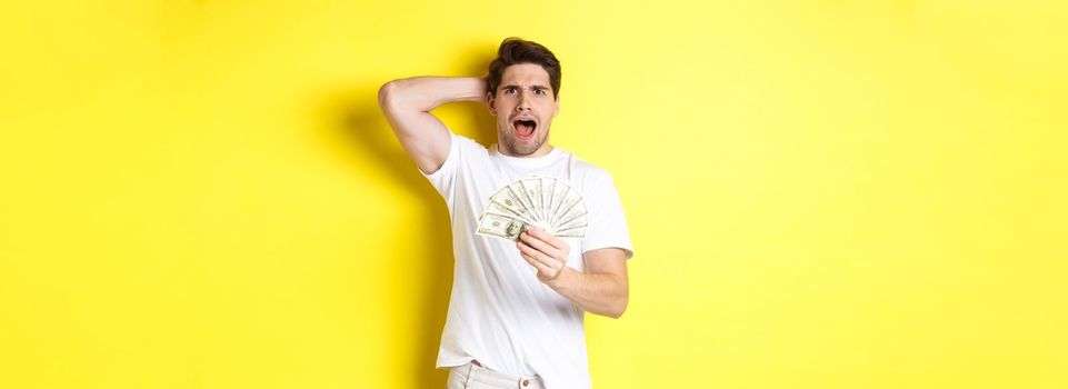 Frustrated man holding money, shouting and panicking, standing over yellow background.