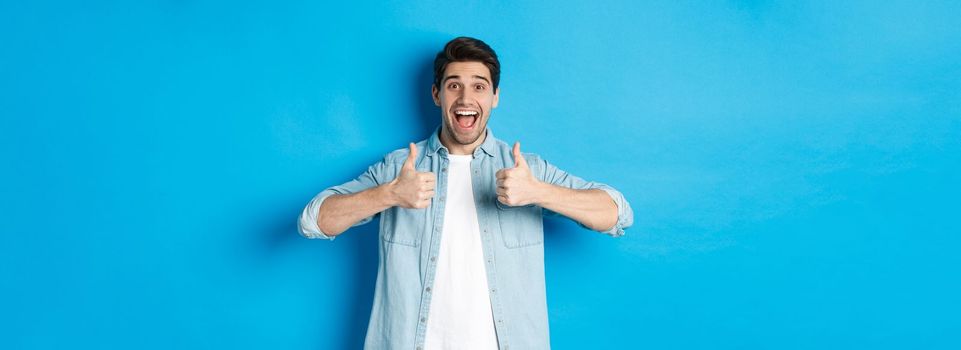 Smiling adult man showing thumbs up with excited face, like something awesome, approving product, standing against blue background.