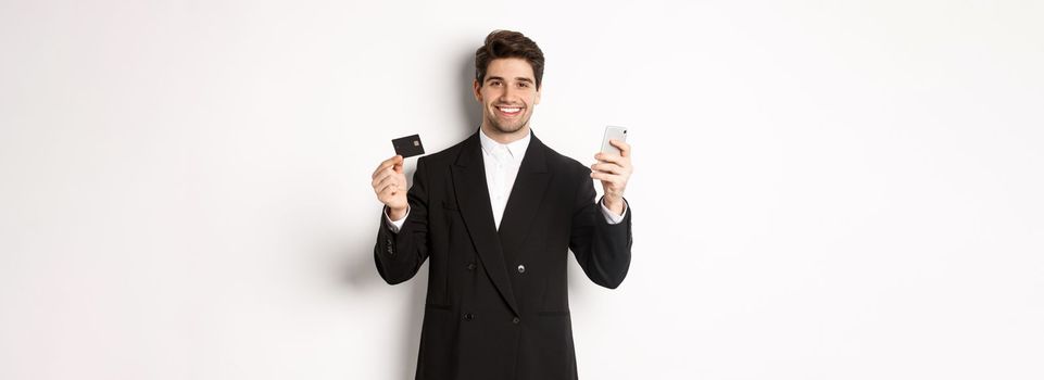 Handsome businessman in black suit smiling, showing credit card and money, standing against white background.