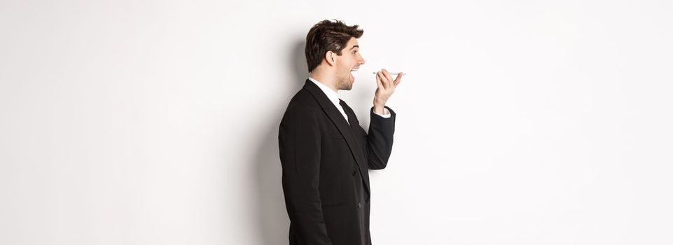 Profile shot of handsome businessman in black suit talking on speakerphone, smiling and looking happy, recording voice message, standing over white background.