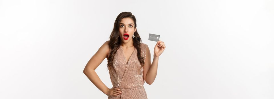 Holidays. christmas party and shopping concept. Amazed attractive woman with red lips, luxury dress, showing credit card and staring at camera, standing over white background.
