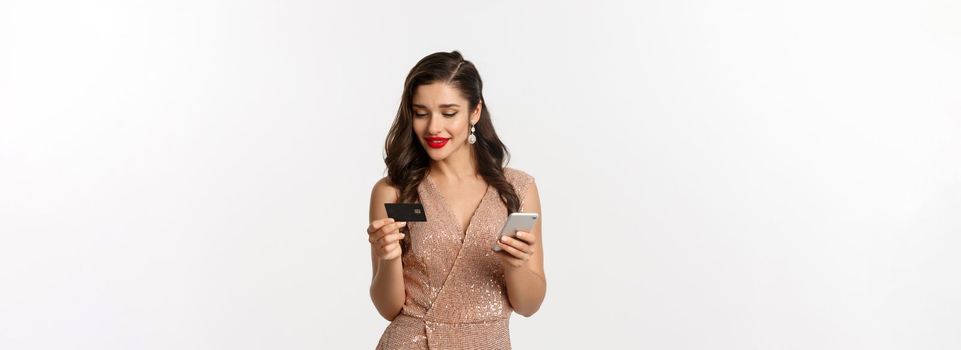 Online shopping and christmas concept. Elegant young woman paying for internet purchase with smartphone and credit card, standing in luxury dress over white background.