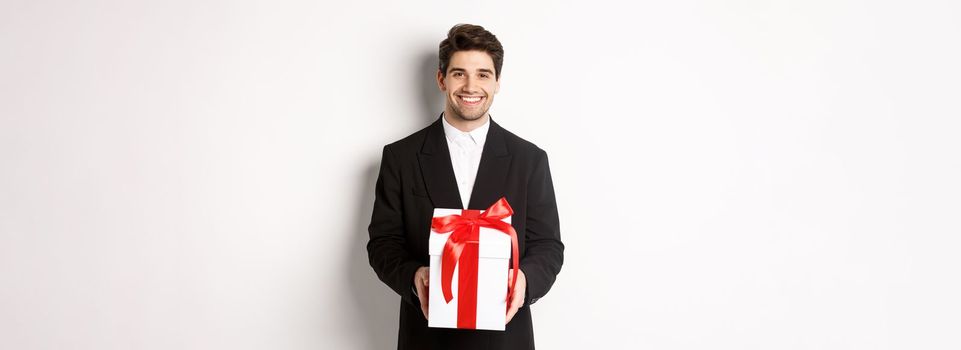 Concept of christmas holidays, celebration and lifestyle. Handsome man in black suit, have romantic present, holding gift in a box and smiling, standing against white background.