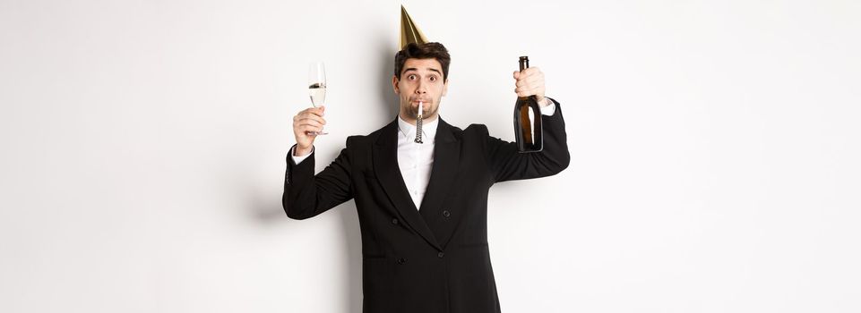 Concept of holidays and lifestyle. Handsome guy celebrating birthday, blowing party whistle and holding champagne, saying a toast, standing in suit over white background.