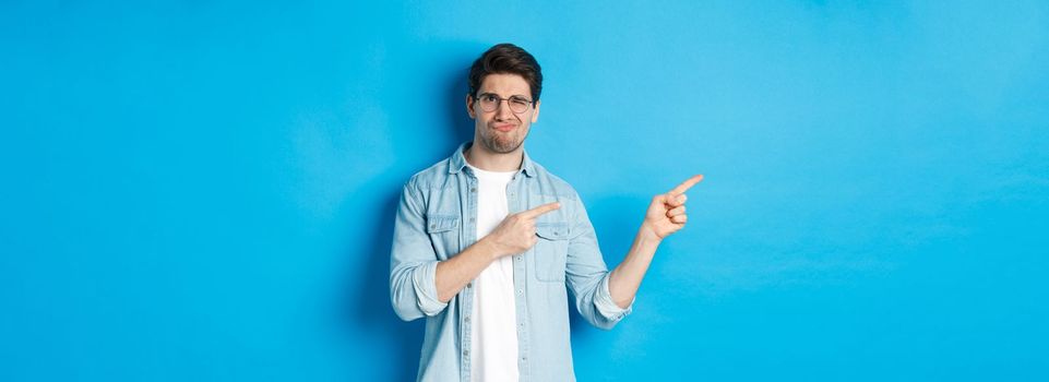 Displeased and skeptical male model in glasses, pointing fingers left at something bad, showing awful advertisement, standing over blue background.