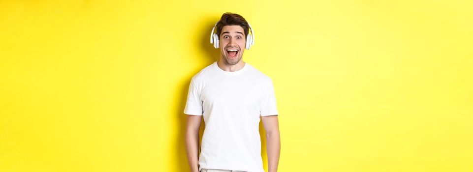 Man in headphones looking surprised, standing against yellow background in white outfit.