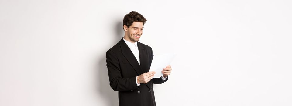 Image of attractive businessman in black suit, reading document and smiling, working on report, standing against white background.