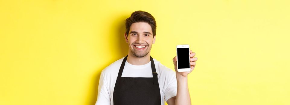 Close-up of handsome waiter in black apron showing smartphone screen, recommending app, standing over yellow background.