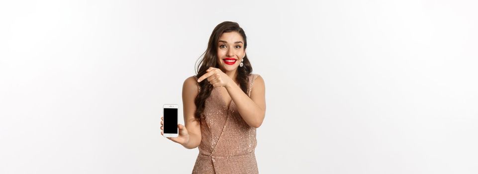 Online shopping. Excited young woman with red lips, elegant dress, pointing finger at mobile screen, showing something, standing over white background.