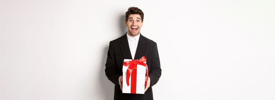 Concept of christmas holidays, celebration and lifestyle. Image of handsome guy in black suit looking excited, have a gift, standing against white background.