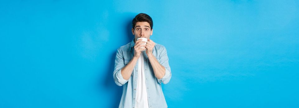 Man looking excited and sipping tea or coffee from white mug, standing over blue background.