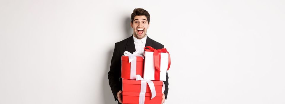 Concept of christmas holidays, celebration and lifestyle. Image of handsome amazed guy in suit, holding new year presents and smiling, standing against white background.