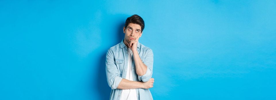 Annoyed adult man rolling eyes and looking bored, standing indifferent against blue background in casual outfit.
