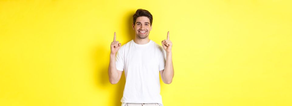 Handsome guy in white t-shirt pointing fingers up, showing shopping offers, standing over yellow background.