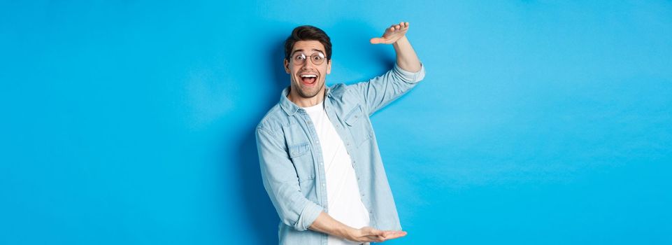 Happy man in glasses showing big size object, shaping large box, standing over blue background and smiling.