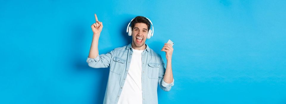 Joyful handsome man dancing with smartphone, listening music in headphones and pointing finger up, standing over blue background.