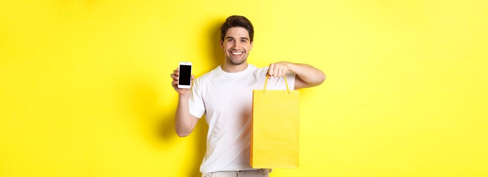 Concept of mobile banking and cashback. Young happy guy holding shopping bag and showing smartphone screen, yellow background.
