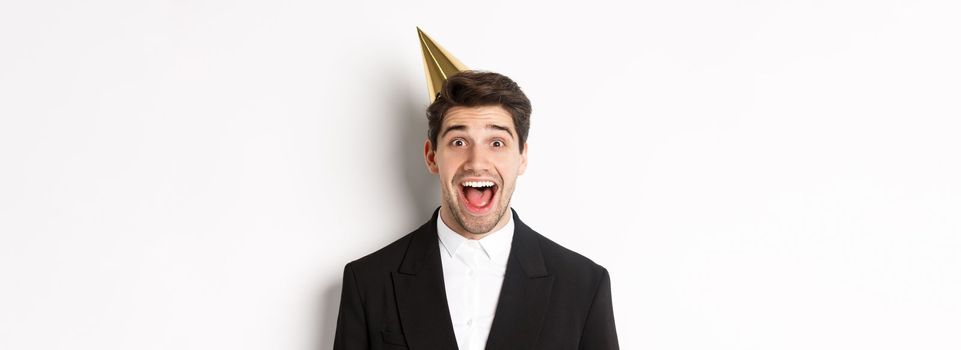 Close-up of excited handsome guy in trendy suit and party hat, looking amazed, celebrating winter holidays, standing against white background.