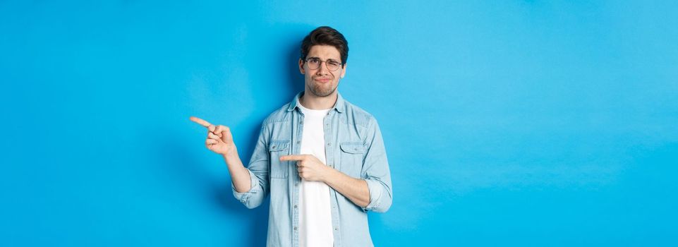 Disappointed young man in glasses pointing fingers right at copy space, showing promo banner and smirking dissatisfied, judging bad product, standing over blue background.