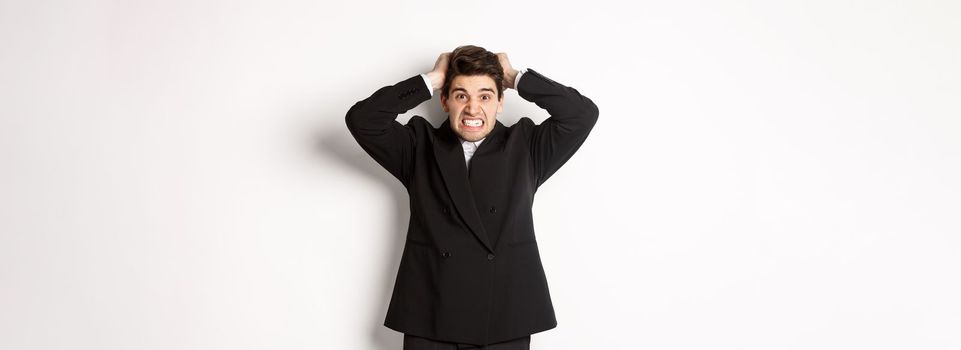 Image of frustrated and angry businessman in black suit, ripping hair on head and grimacing mad, standing tensed against white background.