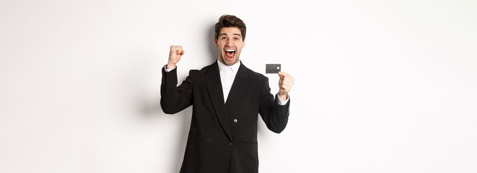Portrait of excited handsome businessman in suit, rejoicing and showing credit card, standing against white background.