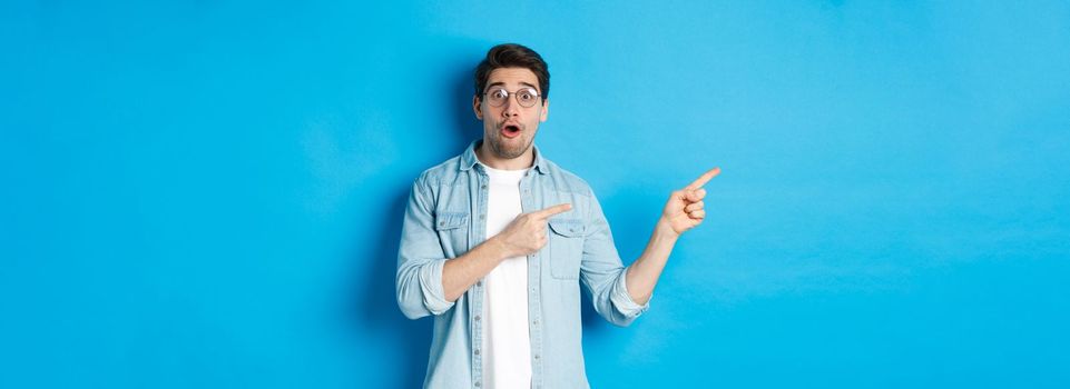 Surprised man in glasses pointing fingers left at copy space, asking question about product, looking intrigued, standing over blue background.