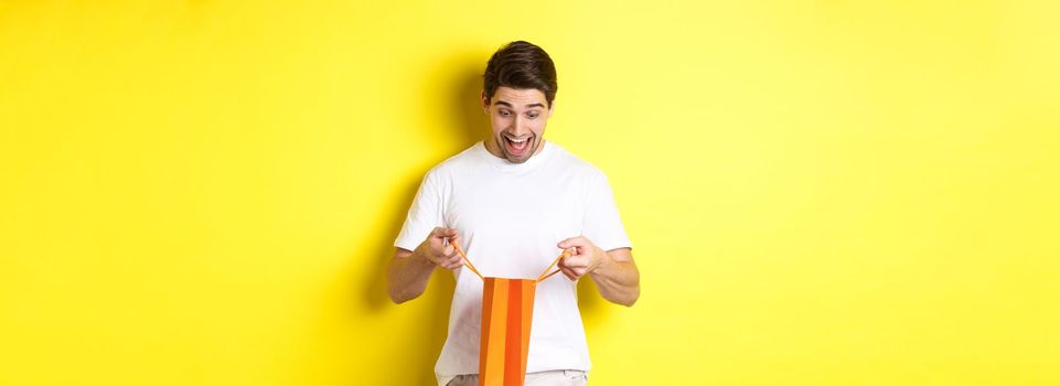 Excited guy open bag with gift, looking inside with amazement and happy face, standing against yellow background.