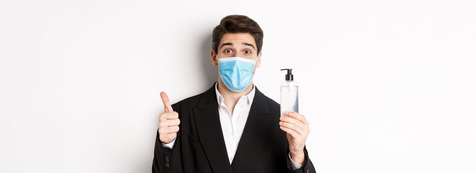 Concept of covid-19, business and social distancing. Close-up of satisfied handsome man in suit and medical mask, showing thumb-up and hand sanitizer, standing against white background.