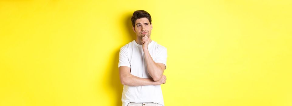 Portrait of young male model thinking, looking at upper left corner and making choice, standing near copy space, yellow background.