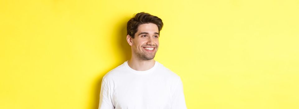 Close-up of attractive bearded man in white t-shirt smiling, looking left at copy space, standing against yellow background.