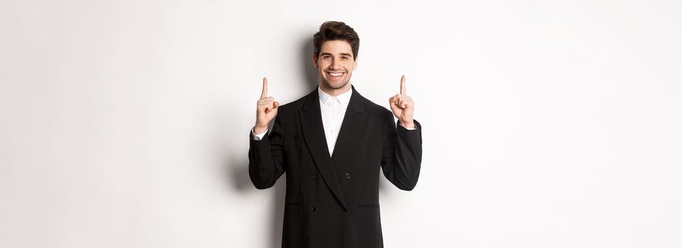 Portrait of attractive caucasian man in stylish black suit, pointing fingers up and smiling, showing christmas advertisement, standing over white background.