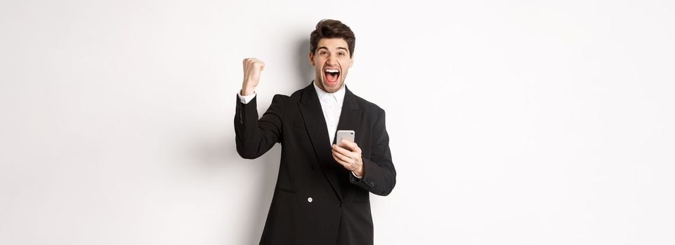 Portrait of happy handsome man in suit, rejoicing, achieve goal on mobile app, raising fist up and shouting yes, holding smartphone, standing against white background.