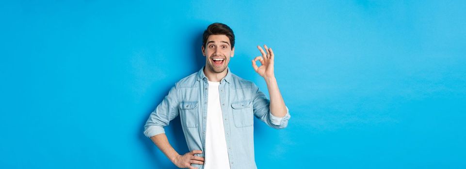 Smiling happy man showing ok sign and looking pleased, approving something good, standing against blue background.