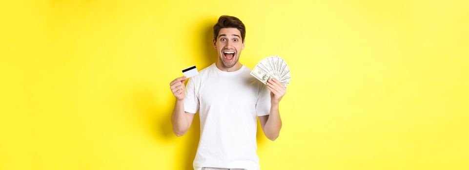 Excited man ready for black friday shopping, holding money and credit card, standing over yellow background.