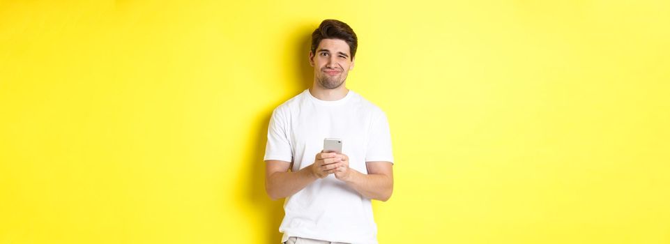 Displeased and reluctant man grimacing, being unamused by message on smartphone, standing over yellow background.