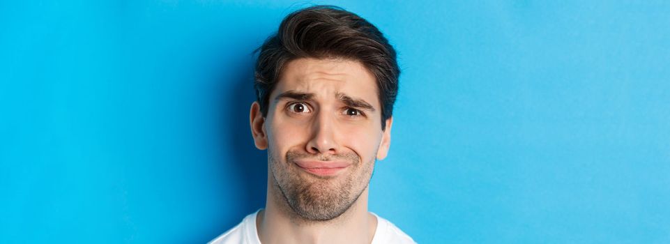 Head shot of skeptical and disappointed man, grimacing complicated, standing against blue background.