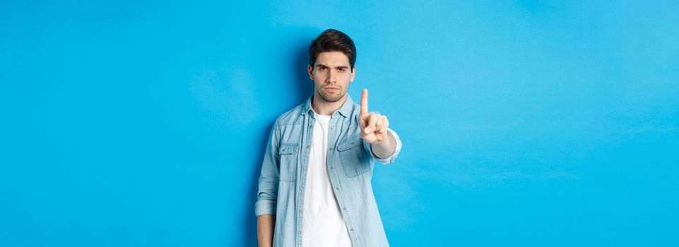 Angry 25 years old man shaking finger in disapproval sign, frowning disappointed, forbid something bad, telling no, standing against blue background.