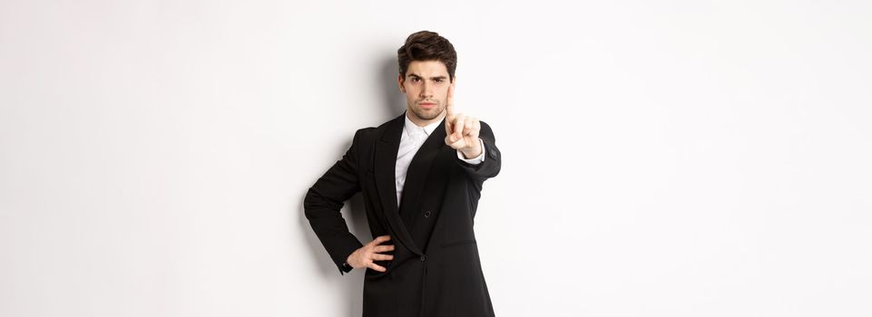 Portrait of serious handsome man in business suit, showing one finger to prohibit or decline something, telling to stop, disagree with you, standing over white background.