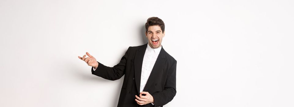 Portrait of happy man dancing at party, playing on invisible guitar and laughing, standing in black suit against white background.