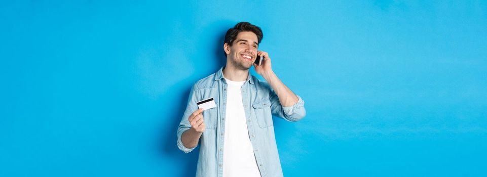 Smiling man call bank support, talking on mobile phone and holding credit card, standing against blue background.