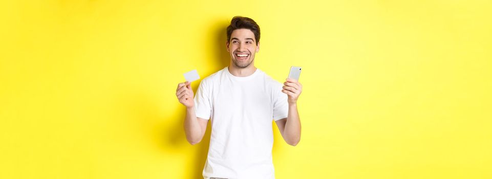 Happy man thinking about shopping, holding credit card and smartphone, smiling pleased, standing over yellow background.